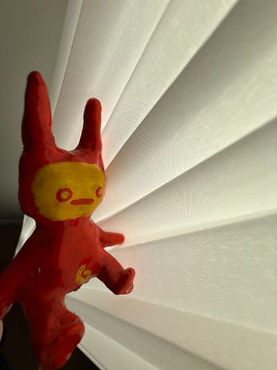 A close-up shows a red, seemingly handcrafted, animal figurine lying against a backdrop of white, pleated window blinds. The animal has a yellow patch on its back, marked with a red exclamation point and circular details. Its limbs are outstretched, giving the impression that the animal is resting or has fallen. The red paint on the animal has a glossy sheen, highlighting its texture.