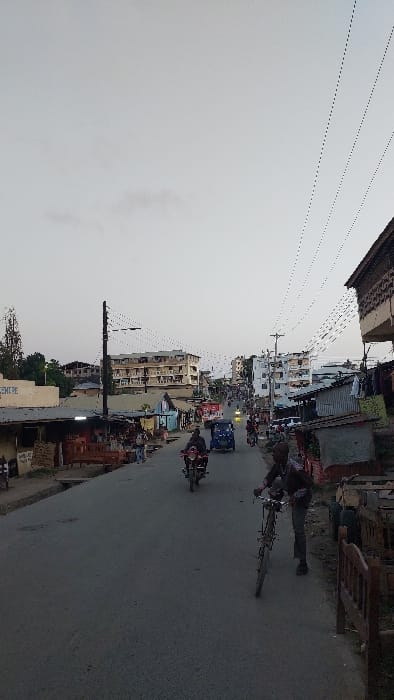 This image depicts a street scene in Magogoni ward, Kenya, captured under an overcast sky. The time of day appears to be either early morning or late afternoon due to the subdued, diffused light.
The primary subject in the foreground, on the right side of the paved road, is a male individual, likely an adult, dressed in a dark long-sleeved shirt and dark trousers, pushing a dark-colored bicycle. The road surface is asphalt, showing signs of wear and minor imperfections. Several vehicles are present on the road: at least two motorcycles, commonly known as boda-bodas, with riders are visible, one red and closer to the foreground. Further up the street, a blue three-wheeled auto-rickshaw (tuk-tuk) is observed, along with other distant vehicles, including what appears to be a car.
On the left side of the road, a series of single-story and low-rise commercial structures or market stalls are visible. One cream-colored building prominently displays a partial sign reading "CENTRE." Wooden benches and various goods are arranged outside these establishments. A utility pole with multiple overhead electrical wires is positioned on the left sidewalk. In the distance on the left, some trees are discernible.
The right side of the road features a more immediate, elevated structure constructed from wood and corrugated metal, with clothes hanging from an upper section. Further down the street, both sides are lined with a mixture of multi-story buildings, some appearing residential or commercial, alongside smaller, less formal structures, including some with corrugated metal roofs. The general architectural style is varied, with some buildings showing signs of age or partial construction.
Numerous thin black electrical wires crisscross the sky, extending between utility poles throughout the visible streetscape, indicating a dense urban or semi-urban infrastructure. The sky itself is a uniform light gray, providing a soft, consistent illumination to the entire scene.