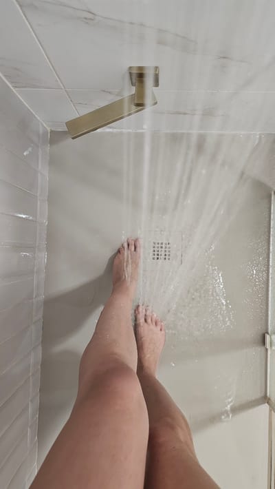 The image shows a person's legs and feet standing in a shower, with water spraying from a modern showerhead onto the floor. The shower has white tiled walls and a light-colored floor with a drain. The person is wearing no clothing. The image is taken from a top-down perspective, focusing on the lower half of the body. There are no discernible elements that would place this image in María De Huerva, Spain.