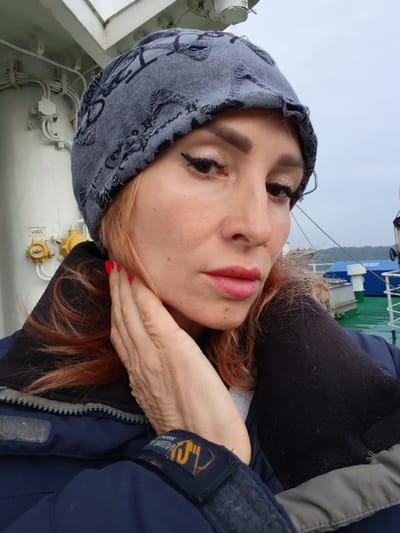 A woman with blonde hair, wearing a grey beanie and a dark blue jacket, poses for a selfie on the deck of a ferry. She has red nail polish on her fingers, which are resting on her cheek. The background shows the railing of the ferry, some industrial equipment, and a cloudy sky. The image was taken on the ferry between Kurikaevo and Kosa in Russia.