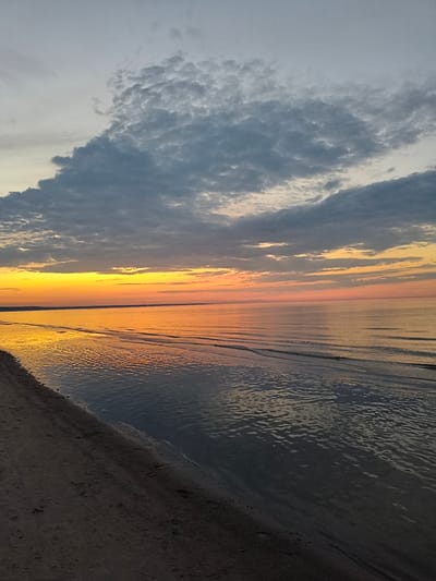 This is a vertical, eye-level, full-shot photograph of a beach at dusk in Apšuciems, Latvia. The sky is filled with dramatic, grey, textured clouds that stretch across the upper half of the frame. Below the clouds, a vibrant band of orange and yellow light emanates from the horizon, indicating the setting sun. The calm sea reflects the colors of the sky, with gentle waves lapping at the shore. The foreground is dominated by a sandy beach, which is dark and appears damp near the water's edge. There are no people or visible man-made objects in the frame.