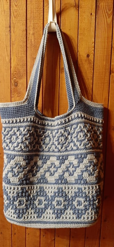 A crocheted tote bag, in a blue and white pattern, hangs from a white hook against a background of vertical wooden planks. The bag has two straps and a textured pattern of geometric shapes. The wooden planks appear to be part of an interior wall. The image does not contain any information about the city of Былина Снт, Russia.