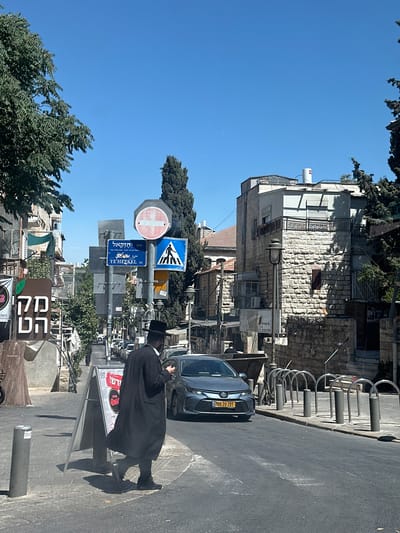 This image depicts a street scene in al-Eizariya, Palestinian Territories, under a clear blue sky, suggesting a bright, sunny day. The orientation of the photo is vertical. A person dressed in traditional black Hasidic Jewish attire, including a dark coat and hat, walks away from the camera on the right side of the frame, casting a long shadow on the paved street.
In the background, a dark blue car with an Israeli license plate (760-20-201) is parked on the road. A row of silver bicycle racks lines the sidewalk. The architecture visible includes buildings constructed with stone and lighter-colored facades. Several street signs are prominent: one is a blue and white pedestrian crossing sign, another is a circular sign with a red border and a white cross, and a blue street sign indicates "YE'HEZKEL ST." in English and Hebrew, along with Arabic text.
Further down the street, there are trees and more buildings. In the foreground on the lower right, a section of a brown sign with Hebrew lettering and a green leaf graphic is visible, possibly indicating a business. Another sign displays the word "פיצות" (pizzas) in Hebrew. The overall impression is of a busy urban street with a mix of architectural styles and cultural elements. The lighting suggests it is daytime, likely late morning or afternoon due to the distinct shadows.