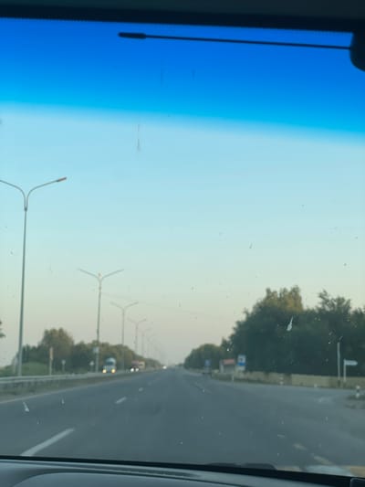 The image depicts a view from inside a vehicle traveling on a highway in the vicinity of Zhyemkum, Kazakhstan. The perspective is from the driver's seat, looking forward through the windshield. The highway is paved and has multiple lanes, with white and yellow lines marking the traffic lanes. Streetlights are visible along the sides of the road, and trees line the median and the shoulders. The sky transitions from a deep blue at the top to a lighter hue towards the horizon, suggesting it is either early morning or late afternoon. A few vehicles, including a white truck and some cars, are visible in the distance. The windshield has several smudges and specks, indicating it is not perfectly clean. The overall scene is consistent with travel on a rural or suburban highway.