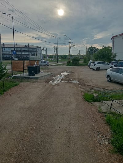 The image captures an overcast sky with the sun peeking through, casting a soft light. In the foreground, a dirt road shows signs of recent rainfall, with several puddles and tire tracks indicating vehicle movement. To the left, two large black trash bins sit on a concrete pad, partially filled with trash bags. Behind the bins, a wooden fence encloses an area, and a "P" parking sign with an arrow points to the left.
Further back, a multi-story building with numerous windows and a dark roof dominates the left side of the frame. A few cars are parked in front of it. The road continues to a parking lot on the right, where several vehicles are visible, including a silver Toyota Prius and a darker SUV. Trees and foliage line the background, suggesting a semi-urban or suburban setting. The scene is consistent with an outdoor area in a settlement, likely a residential or commercial zone, potentially in Chigiri, Russia, given the architectural style and general environment. There are no people visible in the image.