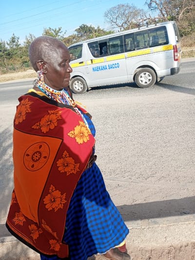 The image displays an outdoor scene in Ildamat ward, Kenya, under bright, clear daylight. In the foreground, an elderly dark-skinned female, with a shaven head and visible facial wrinkles, stands facing right with a slight downward gaze. She is wearing multiple multicolored beaded necklaces and large, intricate beaded earrings. Her attire includes a blue and black checkered garment visible from the waist down, overlaid by a red, orange, and black patterned shawl featuring orange hibiscus-like flowers and a circular motif. A dark belt is present at her waist. In the mid-ground, a silver minivan with a roof rack is positioned on a paved road. The vehicle bears a yellow horizontal stripe and the text "NAEKANA SACCO" in blue capital letters on its side. The background consists of sparse green vegetation, several trees, and a clear blue sky. Distinct shadows are cast on the light gray ground and paved road.