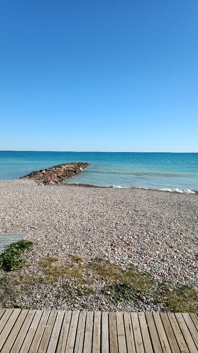 The image depicts a clear, sunny daytime scene at a pebble beach in El Grau De Moncofa, Spain. The foreground features a light brown wooden plank walkway, positioned horizontally at the bottom of the frame, alongside a small section of grey gridded pavement on the far left. Immediately beyond these structures, patches of green and yellowed vegetation grow amidst a vast expanse of light-colored, varied-sized pebbles that constitute the entire beach.
Approximately centered in the midground, a dark-colored rock jetty, constructed from large, irregular stones, extends from the left side of the shore into the calm, turquoise-blue sea. Small, white-capped waves are breaking gently near the shoreline, particularly to the right of the jetty. The sea water exhibits a gradual color change, transitioning from a lighter greenish-blue near the immediate shore to a deeper, more saturated blue-green further out towards the horizon. The horizon line is straight and level. The top portion of the image, approximately 40%, is dominated by a clear, unblemished vibrant blue sky, devoid of any clouds. No individuals, animals, or human activity are visible within the scene.