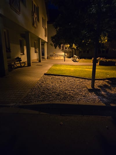The image shows a nighttime scene outside a building in Kostelec na Hané, Czechia. The building, which appears to be a residential or commercial property, is illuminated by artificial light, casting long shadows. In the foreground, a paved walkway made of interlocking pavers leads towards the building. Adjacent to the walkway is a gravel bed, bordered by a curb. Beyond the gravel bed is a patch of green grass. A tree with dark foliage stands in the middle ground, its trunk illuminated by the light. In the background, a wooden bench is visible on the paved area, and further back, flower boxes are present. The overall impression is one of quiet, dimly lit surroundings.
