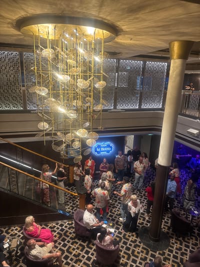 The image depicts an interior scene within what appears to be a cruise ship or a similar entertainment venue. A large, ornate golden chandelier hangs from the ceiling. A group of people are gathered on a lower level, some dancing, others standing and talking. A band is visible on the right, playing instruments. A sign that reads "Le Bistro" is illuminated on the wall. There is a staircase on the left with people standing on it. Several individuals are seated in purple chairs in the foreground. The flooring is patterned with black, gold, and white geometric shapes. The lighting is warm and ambient, creating a festive atmosphere. The location of Motril, Spain is not discernible from the image.