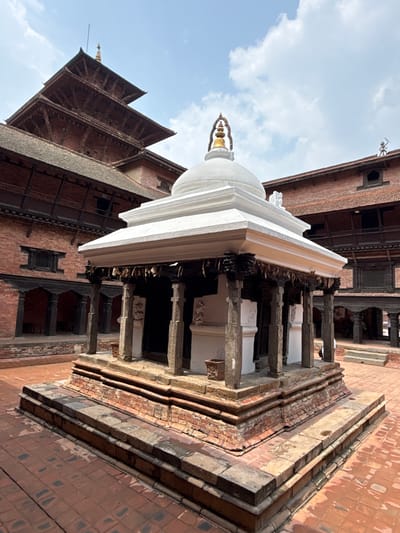 The image captures a low-angle view of a structure in Lalitpur, Nepal. The primary subject is a white stupa with a gold-colored finial, positioned atop a multi-tiered platform. The platform features a series of dark wooden pillars and beams, partially enclosing the base of the stupa. The platform's exterior is constructed of red brick. Behind the stupa, there is a multi-story building with a series of steeply pitched roofs. The building's exterior is characterized by dark wood and latticed windows. The sky is visible in the upper left, with a mix of blue and white clouds. The scene appears to be taken in an open-air courtyard.