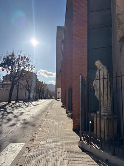 The image displays an outdoor scene of a street on a sunny day. The perspective is from a slightly elevated position, looking down and along the length of the street. The sun is bright and low in the sky, positioned in the upper left quadrant of the frame, casting long shadows and creating a visible lens flare. The sky is clear blue with a few white, fluffy clouds visible towards the upper right.
To the left of the image, a building with red-brown brickwork dominates the foreground. Above the brick section, there's a darker, possibly gray or green, flat façade or roofline. Adjacent to this brick building, closer to the center, a white or light-colored statue of a draped figure stands behind a dark metal fence or railing. The statue appears to be a religious figure, possibly with its head bowed. The fence has vertical bars and some ornamental elements at its top.
A paved street or road extends from the middle-left of the image towards the background, gently curving to the right. On the right side of the street, a sidewalk with light-colored, square tiles runs parallel to the road. The street and sidewalk are well-lit, and the strong sunlight casts prominent, elongated shadows of trees across the road and part of the sidewalk.
Along the right side of the street, a line of trees with bare or sparsely leaved branches suggests either autumn or winter. Beyond these trees, further down the street, additional buildings can be seen. In the distant background, mountains or hills are visible against the horizon.
The overall impression is one of a quiet street on a bright day. There is no visible activity or people. No legible text is apparent in the image.
