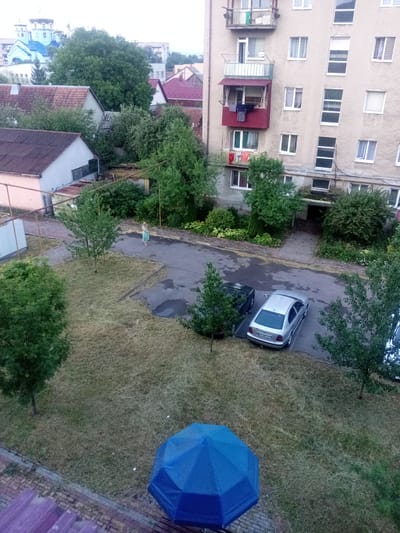 This is a high-angle, vertical medium shot of an outdoor scene in what appears to be a residential area. The focus is on an apartment building and the surrounding courtyard.
In the foreground, a large, vibrant blue umbrella dominates the lower portion of the image. Behind it, a patch of dry, mown grass covers a sloped area. Further back, a paved area with visible tire marks suggests a parking lot or driveway. A silver sedan is parked here, its license plate partially visible. A dark-colored car is parked to its left. A young woman in a light-colored dress is walking on the paved area, facing away from the camera.
The middle ground features a building with a reddish-brown tiled roof and a pale pink facade. Beyond this, several other residential buildings are visible, some with red roofs and others with lighter colors. Trees, some with full green canopies and others with sparser foliage, are scattered throughout the scene.
The background consists of more buildings and trees, suggesting an urban or suburban environment. A distinct blue-domed structure with white architectural details is visible in the distance.
The lighting suggests it is daytime, possibly late afternoon or early evening. The presence of the blue umbrella and the slightly overcast sky hint at possible rain or damp weather, though the grass is visibly dry and mown. The overall scene conveys a sense of everyday life in a residential complex, with elements of urban infrastructure and natural vegetation. No distinct emotions are conveyed, but the scene is peaceful and ordinary. The license plate on the silver car appears to read "EA0395 TE".