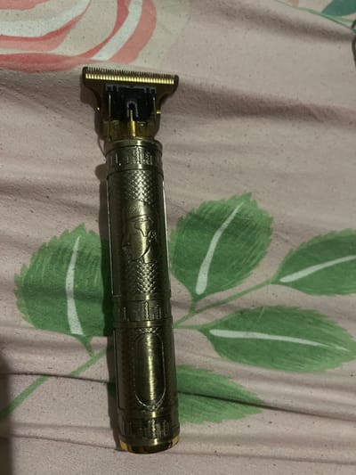 A gold-toned electric trimmer with ornate detailing is placed on a pink and white striped fabric with green leaf patterns. The trimmer features a metallic textured body and a gold-colored blade head. The fabric appears to be a bedsheet or similar textile. The image does not provide any information about the city of Astoņi, Latvia.