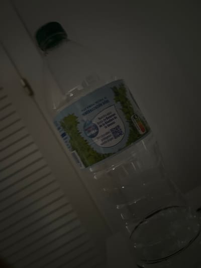 The image shows a plastic water bottle with a green cap, lying on its side. The bottle has a label with text and graphics. The label features an illustration of trees and a landscape, along with nutritional information labeled "NUTRI-SCORE". There is also a QR code. Part of the text on the label is visible and reads "AGUA MINERAL NATURAL DE MINERALIZACIÓN DÉBIL" and "Nuestra agua RECORECTA brota de Reservas NATURAL de la Biosfera de La Unesco. Descubre cómo protegemos nuestro origen:". The image is dark, with the bottle being the primary illuminated subject. The background appears to be a white surface with vertical lines, possibly a refrigerator or a similar appliance. The lighting suggests it might be indoors. The overall scene is a close-up of the water bottle, angled from the bottom left to the top right of the frame.