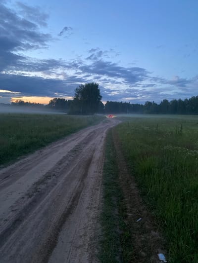 This is a vertical, full-shot photograph of a dirt road in a rural area at dusk. The road is in the foreground and leads into the distance, curving to the left. It is flanked by a grassy field on either side, with some wildflowers visible in the field to the right. In the mid-ground, there is a line of trees and bushes, with a patch of fog obscuring the ground in the distance to the left. A single car with its red taillights illuminated is visible on the road in the distance, just before the trees. The sky is a deep blue with scattered clouds, and the horizon shows a faint orange glow from the setting sun. The scene appears to be peaceful and serene. The image was taken in the city of Yablonnoe, Russia.