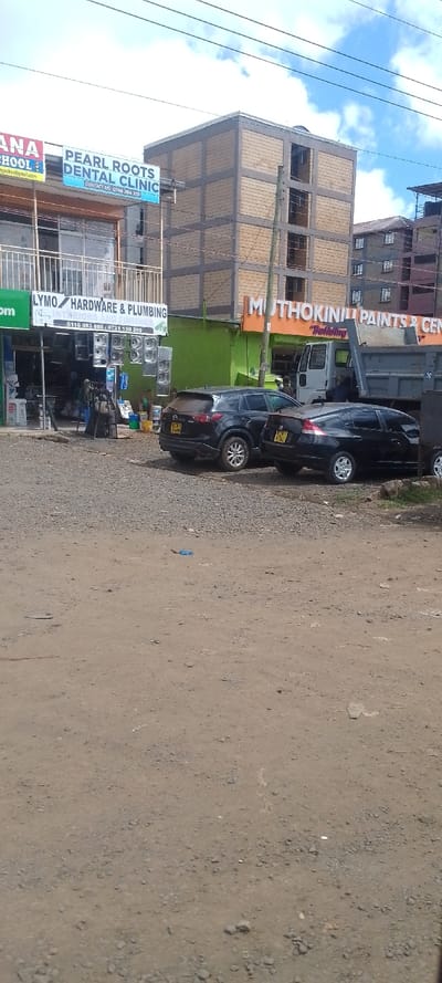 The image displays an outdoor scene in Oloolua ward, Kenya, under a partly cloudy sky. The foreground consists of an unpaved, reddish-brown dirt and gravel surface, with a small blue object resting on the ground in the left-center.

In the midground, from left to right, are commercial buildings and vehicles. A multi-story building on the left features a sign for "PEARL ROOTS DENTAL CLINIC" (contact: 0748 704 333) and below it, "LYMO HARDWARE & PLUMBING" (contact: 0116 083 888 / 0721 150 299), which has numerous stainless steel sinks displayed vertically outside. Two black passenger vehicles are parked in front of the businesses: a Mazda CX-5 SUV (license plate KFN 464W) and a Honda Insight sedan (license plate KFN 976L), both facing away from the viewer. Behind these cars, a white dump truck with a blue/grey bed is partially visible, facing right. To the right of the dump truck is a green storefront with an orange sign for "MUTHOKINIU PAINTS & CEMENTS," with "Building" below it. Several indistinct figures are visible near the dump truck and the Muthokiniu Paints storefront.

The background is dominated by a multi-story, unfinished concrete building with light brown paneling and grey structural elements. Additional multi-story buildings, possibly residential, are visible further back on the right. Multiple overhead utility lines traverse the scene, supported by a utility pole situated in front of the unfinished concrete building.