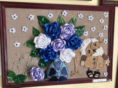 This is a framed, handmade piece of art featuring a bouquet of fabric flowers and a stuffed owl figure. The bouquet, composed of satin ribbons, includes roses in shades of blue, white, and purple, arranged in a blue vase adorned with dark blue and patterned blue stones. Green leaves made of shiny material are interspersed with the flowers. Decorative strings of small, pearl-like beads add a whimsical touch. To the right of the bouquet stands a stuffed owl figure made from burlap and yarn, wearing a knitted hat and coat with button details. The owl has black button eyes and a black beak. Small white fabric flowers with blue centers are scattered throughout the composition. The artwork is presented against a tan, burlap-like background within a dark wooden frame. A small label with text in Cyrillic is visible at the bottom right of the frame. The artwork appears to be displayed on a wall. The location is stated as Ветрынскі сельскі Савет, Belarus.
