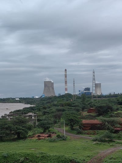 The image depicts a power plant with tall cooling towers and smokestacks against a cloudy sky. The plant is situated near a river with a murky brown color. The foreground is covered in lush green vegetation, including trees and grass. There are also some brick structures visible in the lower right portion of the image. The scene suggests an industrial area adjacent to a natural landscape, likely Fekari, India, given the context provided. There are no people or animals visible in the image, and no obvious actions or interactions are taking place. The environment appears to be somewhat overcast, possibly indicating a rainy or humid day.