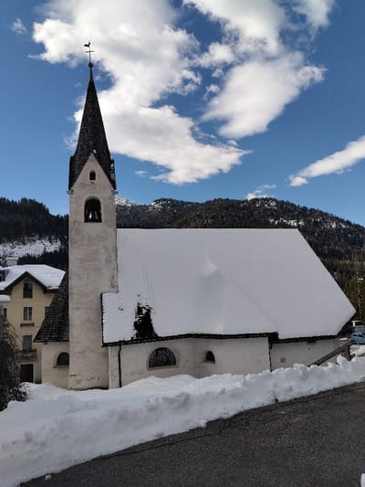 The image displays a white stucco church in Serdes, Italy, during winter conditions. The church's main structure features a steep gable roof entirely covered by a thick layer of white snow. A prominent, slender bell tower rises from the left side of the church, capped by a dark, conical shingle spire that supports a cross and a weathervane. The tower includes an arched belfry opening revealing a bell, and a smaller rectangular window above it. On the visible side facade of the church, two arched windows fitted with dark decorative grilles and one smaller, oval-shaped window are present.

The ground surrounding the church is heavily blanketed in white snow, forming a substantial drift along the edge of a dark asphalt road or pathway in the foreground. To the immediate left of the church, a multi-story building with light yellow walls and a dark, partially snow-covered roof is visible. In the background, dark, densely forested mountains rise under a bright blue sky adorned with scattered white cumulus clouds. Some snow patches are visible on the mountainsides. Several light-colored vehicles are partially discernible in the far right background, parked near the base of the mountains. No individuals or specific actions are depicted within the scene.