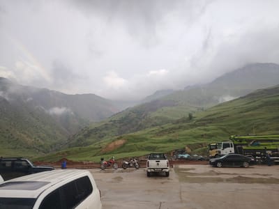 A panoramic view captures a mountain landscape under a cloudy sky, with a faint rainbow visible to the left. The foreground features a paved area with several vehicles, including a white SUV, a black SUV, a white pickup truck, and a black sedan. Two motorcycles are parked near the pickup truck. A large concrete pump truck is parked on the right side of the frame, next to a stack of construction materials. A person in blue clothing is standing near the truck. The terrain is rugged and green, with steep hillsides and a dry riverbed or ravine running through the valley. The weather appears overcast, possibly after rain, given the wet appearance of the pavement and the presence of the rainbow. The scene suggests a construction or infrastructure project in a remote, mountainous region. There is some visible text on the license plate of the white pickup truck: "C 789 KD 01". The location context provided is Farab, Tajikistan.