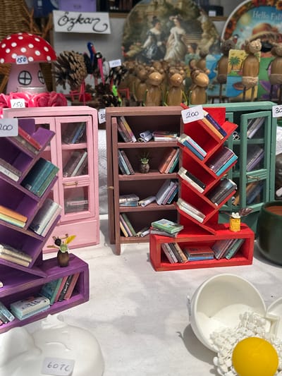 A close-up, eye-level shot displays a collection of miniature decorative items, possibly for sale at a market stall. In the foreground, a white ceramic object, resembling a broken egg or a large decorative shell, is filled with white beads and a single yellow orb. To the left, a purple multi-tiered shelf unit holds miniature books. Behind it, a pink cabinet with glass doors displays more tiny books. A small, brown vase with a delicate yellow flower and two green leaves sits atop the purple shelf. To the right, a red, multi-tiered shelf unit is filled with miniature books. Further back, a green cabinet with glass doors also contains tiny books. In the background, several wooden figurines, resembling bears, stand arranged on a surface. A price tag of "150" is visible on a red and white spotted mushroom decoration. Another price tag reads "150" on a pinecone decoration that has been fashioned into a bird. A sign above the mushroom reads "Özkar". The scene is set against a white tablecloth. There are no people visible in the image. The items appear to be crafts or toys, and the overall impression is one of a charming and detailed display. The presence of the sign "Özkar" and the style of the items suggest this could be a vendor's display at a local market or fair in Kayseri, Turkey, known for its craft markets.