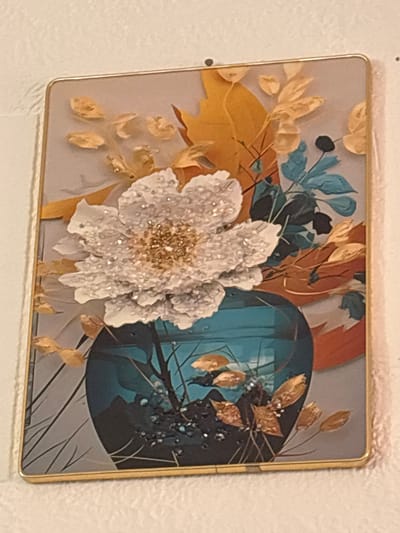 This media file shows a rectangular magnet with a floral art print. The print features a large, white, textured flower with a gold center, surrounded by various botanical elements. These include golden leaves and stems, dark blue and black leaves, and a large, dark blue, circular object in the background that appears to be a vase or an abstract element. The overall style is decorative and somewhat abstract, with a mix of organic and geometric shapes. The magnet has a thin gold border and is attached to a white, textured surface, likely a refrigerator or a similar appliance. No people, text, or specific time of day or weather conditions are visible in the artwork or its immediate surroundings. The location context provided is Zlataritsa, Bulgaria, but there are no visual cues within the image that directly support this location.
