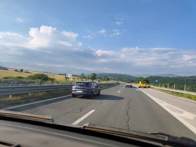 The image captures a view from inside a vehicle traveling on a highway in the vicinity of Dzhurovo, Bulgaria. The highway is a paved, multi-lane road with white lane markings. In the foreground, the dashboard of the vehicle is visible. On the highway, there are several vehicles including a dark gray hatchback in the left lane, a white pickup truck further ahead in the right lane, and a yellow van to its right. The surrounding environment consists of rolling hills with vegetation, fields, and a clear blue sky with scattered white clouds. A metal guardrail separates the highway from the roadside. Distant mountains are visible on the horizon. The overall scene suggests a sunny day with moderate traffic.