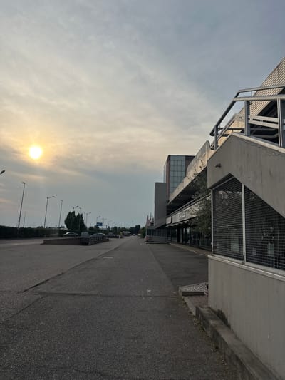 The image depicts an outdoor scene with a bright sun partially obscured by clouds on the left side, casting a soft glow. A paved area, possibly a parking lot or street, dominates the foreground and extends into the distance. On the right side of the frame, a concrete structure with a metal railing and a wire mesh enclosure is visible. Further down the paved area, a multi-story building with a modern facade and glass windows is present, accompanied by trees and streetlights. Several cars are parked or driving in the background. The environment appears to be an urban or commercial area in San Martino Buon Albergo, Italy, under a hazy sky. There are no clearly discernible individuals in the frame.
