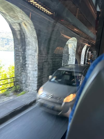 The image was taken from inside a bus traveling on a road, likely in Mandello del Lario, Italy (latitude: 45.944, longitude: 9.309). The bus is traveling under a series of stone arches. A grey car is visible in the foreground, partially obscured by the bus's window, traveling in the same direction as the bus. The car's headlights are visible. The arches are made of rough, grey stone and are partially covered with white paint. The road is paved and appears to be a two-lane road. The bus window is partially obscured by a blue seat. Green foliage is visible through the window, suggesting the road may be near a body of water. The bus is moving, and the image is blurred. The bus is moving quickly, as the car in the foreground is blurry. The scene appears to be lit with natural sunlight. A portion of the bus's ceiling is visible in the upper left of the frame, including a red indicator light and a series of fluorescent lights. The bus appears to be an older model.
