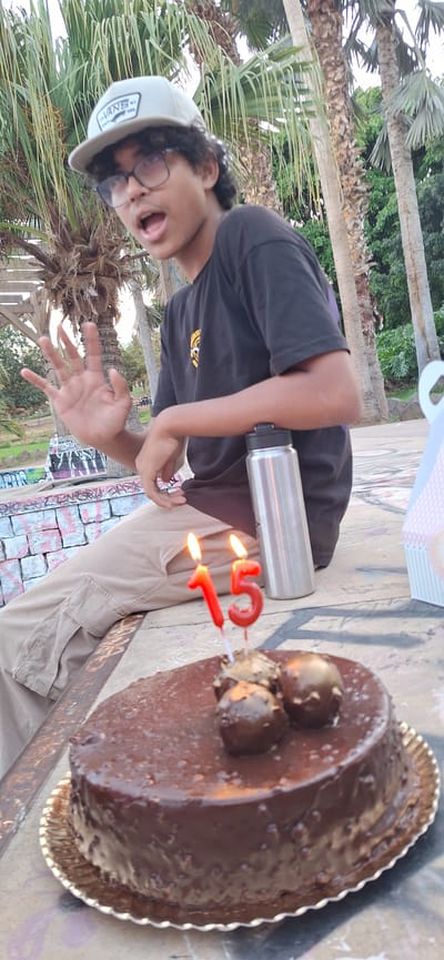 A person is celebrating a birthday outdoors in Santa Cruz de Tenerife, Spain. The subject is a young individual wearing a dark T-shirt with a yellow design, khaki pants, a Vans cap, and glasses. The person has a water bottle and is posing with their tongue out, holding up a hand towards the camera. Next to the person is a chocolate cake with a number 51 candle, suggesting they are celebrating their 51st birthday. The setting appears to be a park or outdoor area with palm trees and graffiti-covered surfaces. There's a hint of green vegetation in the background, contributing to the outdoor environment.