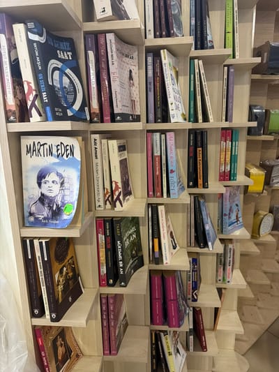 This image depicts a bookshelf filled with books in what appears to be a bookstore or library. The shelves are made of light-colored wood and are arranged in a staggered pattern. The books vary in size and color, with many of their titles visible. On the top shelf, there are books with titles like "ON SEKİZ SAAT" and "MARTIN EDEN". Other visible titles include "ASK TARİFLERİ", "MÜKEMMEL BİR AN", and "ATATÜRK CUMHURİYETİNİN İLK HUTBELERİ". The overall impression is one of a well-stocked collection of literature. The location is identified as Acıpayam, Turkey.