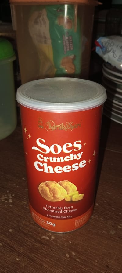 The image features a red cylindrical container of "Soes Crunchy Cheese" by the brand "Kartika Sari," positioned on a wooden surface. The container has a clear plastic lid and displays gold lettering for "Kartika Sari," with prominent white text reading "Soes Crunchy Cheese" accented by stars. An illustration below depicts two golden crunchy pastries and several cheese cubes. Further text identifies the product as "Crunchy Soes Flavoured Cheese" and "Soes Kering Rasa Keju" (Indonesian for Dry Soes Cheese Flavor), with a "Net Weight 50g" indicated.
In the background, a translucent storage container holds a green plastic bag with text visible, including "NCOPP NGETEHI" and "Tuman," suggesting another packaged food item. A green bottle or container is partially visible to the left, and a stack of striped plates is seen on the right. The scene is indoors, characterized by warm, somewhat dim lighting. No specific activity or event is occurring. The visible text and brand names indicate the products are Indonesian, which contradicts the given location context of Aral, Kyrgyzstan.