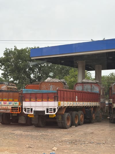 The image depicts a row of parked trucks under a blue canopy structure, likely a covered parking area or a fuel station, in Bethamcherla, India. The trucks are predominantly red and brown, with visible wear and tear. Some trucks have white lettering on their sides, including the phrases "Hello Please Sound Horn Please". The ground is a mixture of dirt and gravel. In the background, lush green trees are visible, suggesting an outdoor setting. The sky is overcast. Two pigeons are perched on the blue canopy. The trucks appear to be commercial vehicles, possibly for transport or logistics. No people are visible in the image.