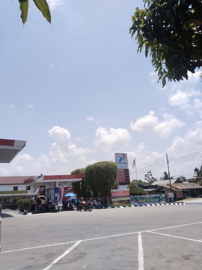 The image depicts an outdoor scene at a Pertamina gas station (SPBU 14 244 479) in Damatutong, Indonesia, under a clear blue sky with scattered white clouds. In the foreground, asphalt pavement with white parking lines is visible. The gas station structure features a red and white canopy, beneath which multiple fuel pumps are positioned. A tall, vertical sign displaying the Pertamina logo and fuel types (Pertamax Turbo 98, Pertamax, Pertalite, Dexlite) stands near the pumps.

Numerous individuals are gathered around the fuel pumps, primarily clustered with several motorcycles. Some people are standing, while at least one person is seated under a blue umbrella. A light-colored truck is partially visible on the far left. An Indonesian flag flies from a pole adjacent to the Pertamina sign. A large banner prominently displays "PT. KANA TAMITA BERSAMA SPBU 14 244 479" and "Idul Fitri." Green foliage from trees is present in the upper corners and borders the station. In the background, residential or commercial buildings with corrugated roofs, along with power lines and poles, are discernible.