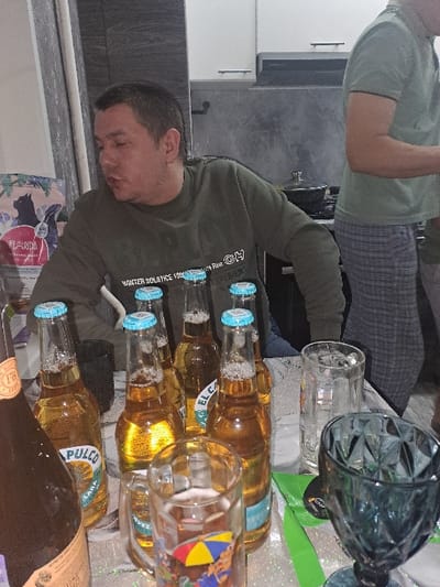 A casual indoor gathering shows a man in a green sweater near multiple bottles of "Pulco" beer, with light blue caps. There are several glasses present, including one with a beach umbrella design and a large, decorative glass container. Another person is visible in a green shirt and gray pants. The setting appears to be a kitchen or dining area with white cabinets and a gray countertop.