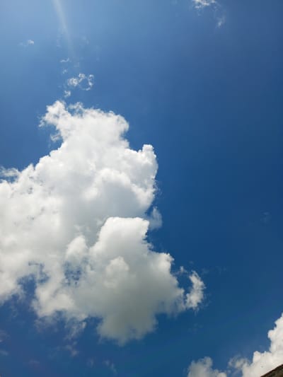 The image is a vertical, eye-level shot of a clear blue sky with several white cumulus clouds. A sunbeam cuts across the upper left quadrant of the image. The lower right corner of the image shows the edge of a dark, angular structure, possibly a building, with three small black birds in flight above it. There are no people visible in the image. The scene depicts a bright, partly cloudy day. The location is identified as Zavyalovo, Russia.