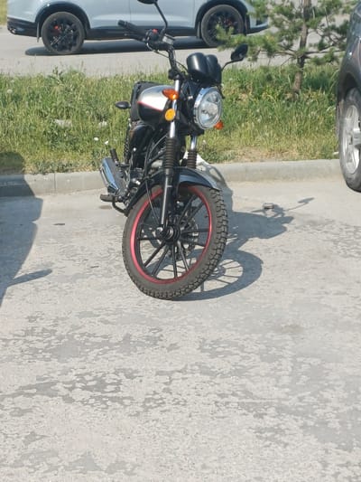A black and silver motorcycle is parked on an asphalt surface in front of a grassy area with a tree. The motorcycle has a chrome exhaust pipe, black handlebars, a round headlight, and a red-rimmed front wheel. In the background, a white SUV is partially visible, and to the right, a portion of a dark-colored vehicle is present. The ground appears weathered and there are shadows cast on it. The scene is outdoors and appears to be a parking area.