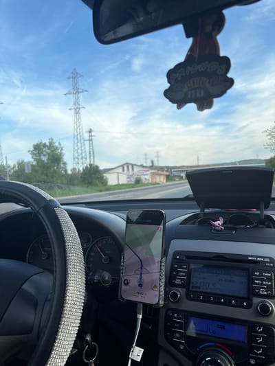 The image depicts the interior of a car, likely in motion. The dashboard is visible, including a car stereo tuned to FM1 96.80MHz, a GPS navigation system running on a smartphone, and a steering wheel with a decorative cover. Through the windshield, power lines and buildings are visible. The GPS indicates a location of approximately 13 km away from the destination "Хан Аспарух," in or near Tulovo, Bulgaria.