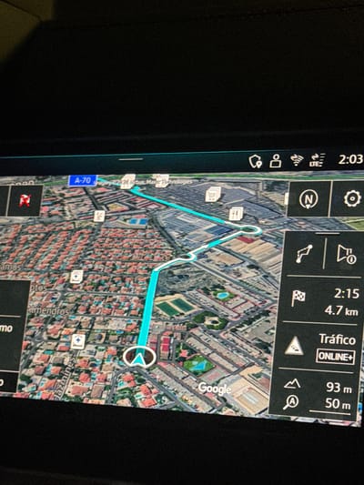 The image shows a car's GPS navigation system displaying a 3D map of a city. The route is highlighted in turquoise. On the right side of the screen, there is a panel with navigation information. It shows a finish line flag icon with "2:15" and "4.7 km" below it, indicating an estimated arrival time and distance. Further down, "Tráfico" (Traffic) is displayed with "ONLINE+" below it, and a traffic sign icon. At the bottom of this panel, there are icons for a mountain peak and a magnifying glass with distances "93 m" and "50 m" next to them. On the left side of the screen, street names like "Almendros" and "Jazmines" are visible. A street sign for "A-70" is also present. The map appears to be of the city of San Vicente Del Raspeig/Sant Vicent Del Raspeig, Spain, with residential areas and some commercial or industrial buildings depicted. The screen is illuminated and appears to be in a dark environment, likely the interior of a vehicle.