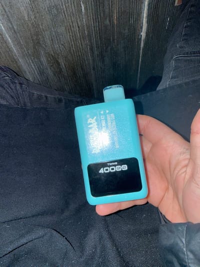 The specific location within Holtzheim, France, is not identifiable from the provided visual evidence. The image, captured from a close-up, elevated perspective, depicts a person's right hand holding a turquoise blue disposable vaping device. The hand, with light skin, is positioned over the person's lap, which is covered by dark-colored fabric, consistent with denim or similar material, showing some lint.

The vaping device is rectangular, with a clear, transparent mouthpiece at the top and a slightly glittery or sparkly finish on its main turquoise body. Text on the device, partially obscured by a bright light reflection, identifies the brand as "NIGBAR" and lists flavors including "PINEAPPLE COLA ICE" and "STRAWBERRY WATERMELON BUBBLE GUM." A black rectangular display panel at the bottom of the device clearly shows "TWINS" and "4000 PUFFS".

No active use of the device, such as vapor production, is visible. The background consists of an indistinct, dark, vertically textured surface, resembling wood paneling or a wall, under what appears to be dim ambient lighting with a strong, localized light source reflecting off the device.