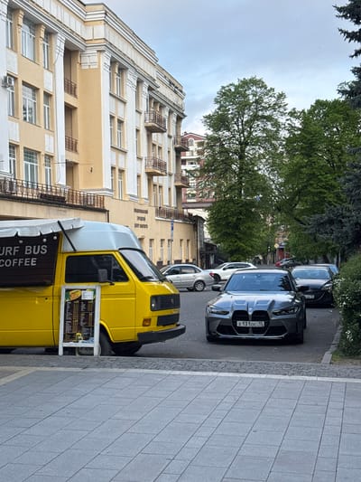 The image shows a street scene in what appears to be Akhieli, Georgia. A yellow van with "SURF BUS COFFEE" signage is parked along the right side of the street, adjacent to a gray tiled sidewalk. Its menu is visible. Parallel to the van, on the road, are several parked cars, including a gray BMW with the license plate "X 131 OC 15". A light-colored, multi-story building with multiple windows and dark iron balconies is on the left. Overhanging trees are present in the center and top sections of the image. The sky is overcast.