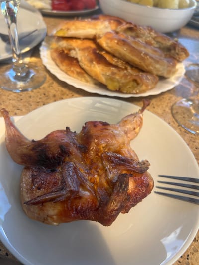 A whole roasted quail is presented on a white plate, accompanied by a fork. In the background, a plate of pastries and a glass are visible. The setting appears to be a table, possibly at a restaurant or a home in the city of Dzerzhinskoye, Moldova.