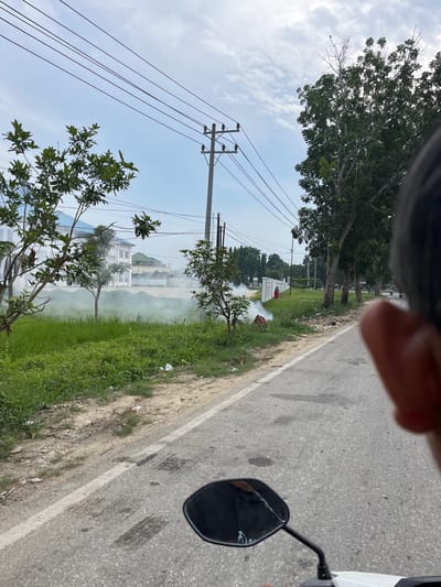 The image depicts a street scene in Tanahterban, Indonesia. In the foreground, a motorcycle's rearview mirror is visible, with a person's finger partially obscuring the view. The road ahead is paved with asphalt and marked with a white line. To the left of the road, a grassy area is interspersed with trees and some buildings, one of which appears to be a white structure with a blue roof. A noticeable plume of white smoke rises from the grassy area, suggesting a small fire or controlled burning is occurring. Power lines are strung across the sky, supported by utility poles. The sky is overcast with scattered clouds. In the background, more trees and buildings can be seen, with several motorcycles further down the road. The overall environment appears to be a mix of residential and utility infrastructure.