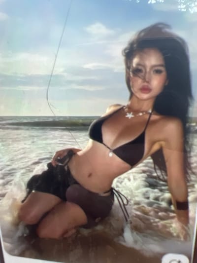 A young woman is kneeling in shallow water near a beach. The woman has long dark hair and is wearing a dark bikini and dark shorts. She is looking towards the viewer with her head slightly tilted. Her body is angled to the left of the frame. The water is foamy and surrounds her legs. The background consists of a body of water with waves and a sky with clouds. There is a thin, vertical line across the image, possibly a scratch on a screen or a thin object in the foreground. The image appears to be a photograph. The location of Kara-Dıykan, Kyrgyzstan is not discernible from the image.
