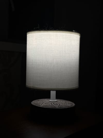 A close-up, eye-level shot features a small, white cylindrical lampshade, softly illuminated from within, sitting on a dark wooden surface. The lampshade has a textured fabric surface and is trimmed with a darker material at the top and bottom. Small wisps of dark material, possibly dust or loose threads, are visible along the top edge of the lampshade. The lampshade is supported by a white rod, which extends down to a decorative, circular base with an intricate, carved pattern. The background is entirely black, indicating a dark room. The image provides no information about the city of Zhovte, Ukraine, or any people, actions, or interactions.