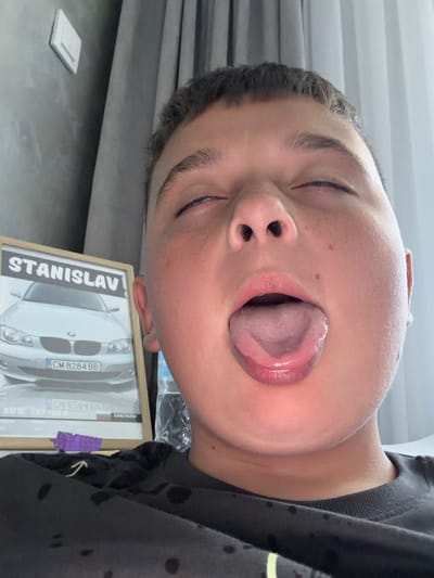 A young boy with his eyes closed and mouth open, tongue extended, is pictured in a close-up selfie. He is wearing a black t-shirt with dark circular stains. To his left, a framed picture displays a silver BMW car with the license plate "CM 8284 BB" and the name "STANISLAV" printed above it. The background consists of grey curtains and a wall-mounted light switch. The scene is likely indoors, possibly in Zmeitsa, Bulgaria, given the context.