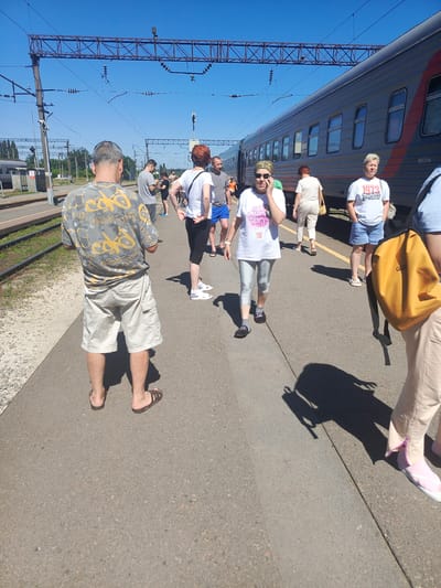 This media file captures a sunny day at a train station in Gryazi, Russia. Several people are gathered on the platform, some looking towards a train that is parked alongside. The train appears to be a passenger train, with a series of windows visible along its side. The platform is paved, and there are train tracks running next to it. Overhead, power lines for the train are strung across metal gantries. The weather is clear and bright, indicated by the strong shadows cast by the people.
The subjects include men and women of various ages, dressed in casual summer clothing. Some are standing, others appear to be walking or waiting. One woman in the foreground is on her phone, looking towards the camera. Another woman to her right is wearing a white t-shirt with text that includes "1973". A man in a grey and yellow patterned t-shirt and beige shorts is standing with his back to the camera. A woman in a white t-shirt and striped pants is walking away from the viewer.
The scene suggests a moment of waiting or transition, possibly for boarding a train or after disembarking. The atmosphere appears relaxed, with no signs of urgency or distress. The presence of a train, tracks, and platform clearly indicates the setting is a railway station. The clear blue sky and bright sunlight point to a time of day likely between late morning and mid-afternoon.