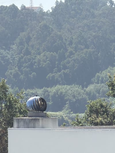 The image shows a rooftop in Gondomar, Portugal, with a landscape of lush, green hills in the background. A distinctive architectural feature, a silver rooftop turbine vent, is prominently visible on the right side of the frame. This vent, designed to exhaust air and provide ventilation, is spherical with curved, metallic blades that create a dynamic, almost sculptural effect.
The background is dominated by a dense forest covering rolling hills. The trees are a vibrant green, suggesting a temperate climate and healthy foliage. The haziness in the air, particularly over the distant hills, implies it could be a warm day with some atmospheric moisture, or perhaps early morning or late afternoon light. The lighting suggests it is daytime, with the sun illuminating the scene and casting shadows, though no specific time can be definitively determined.
In the far left background, hints of human infrastructure are visible: a transmission tower and what appears to be a building with a reddish roof, peeking through the trees. These elements provide a subtle contrast to the natural landscape, indicating the proximity of a populated area to the wilderness.
The scene is devoid of any people, focusing instead on the interplay between man-made structure and nature. The overall mood is serene and natural, with the metallic gleam of the vent adding a touch of modern industrial design against the organic backdrop. There is no visible text in the image.