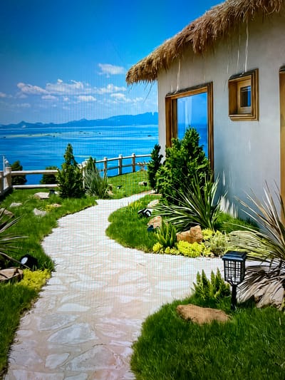 This image depicts a serene outdoor scene in the city of Singgahmata, Indonesia. A winding stone pathway meanders through a lush green lawn, bordered by manicured shrubs and rocks. The pathway leads towards a white building with a thatched roof, featuring a large window that reflects the bright blue sky and the tranquil ocean beyond. In the distance, a calm sea stretches towards a hazy mountain range. A wooden fence runs along the edge of the property, separating it from the water. Several small black solar-powered lanterns are strategically placed along the pathway and in the garden, providing subtle illumination. The overall atmosphere is one of peacefulness and natural beauty.