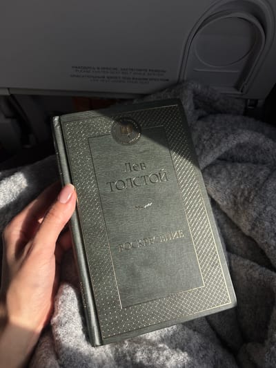 The image shows a book held in a hand, placed on top of a gray, knit sweater. The book is a Russian edition of Leo Tolstoy's "Resurrection" ("Воскресение"). The hand belongs to a person with pale skin and short, manicured nails. The book is dark green, with gold accents. The scene appears to be in an airplane, evidenced by the presence of a partially visible safety instruction panel on the left, indicating "PLEASE FASTEN SEAT BELT WHILE SEATED" and "LIFE VEST UNDER YOUR SEAT" in both Russian and English. Based on the available information, the location can be assumed to be an airplane possibly flying from or towards the city of Yemelyanovo (airport) in Russia. The image suggests someone reading on a flight.